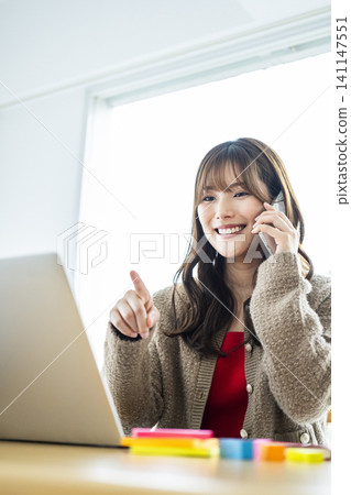 一位年輕女子一邊看著電腦,一邊用智慧型手機開會。 一位年輕女子一邊看著電腦,一邊用智慧型手機開會。 141147551