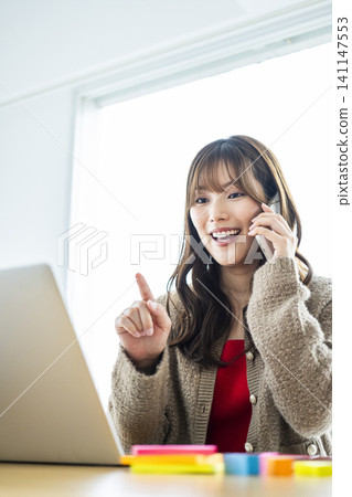 一位年輕女子一邊看著電腦,一邊用智慧型手機開會。 一位年輕女子一邊看著電腦,一邊用智慧型手機開會。 141147553