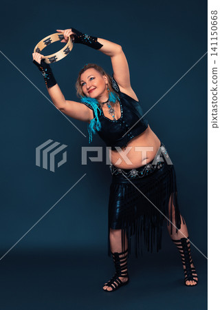 Smiling female performer engages in captivating percussive dance, her movements accompanied by rhythmic striking of tambourine. Caucasian female dancer attired in black handmade performance costume Smiling female performer engages in captivating percussive dance, her movements accompanied by rhythmic striking of tambourine. Caucasian female dancer attired in black handmade performance costume 141150668