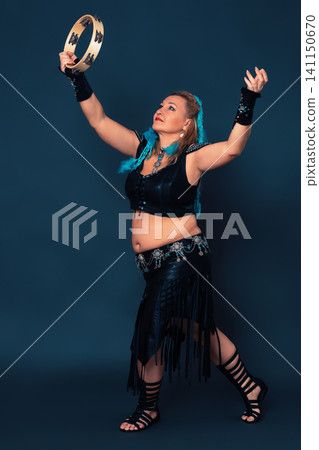 Confidence woman dancing with tambourine, contributing to dynamic ambiance. Curvy female dancer is dressed in black, handmade costume, which includes leather top and skirt, bracers, gladiator boots Confidence woman dancing with tambourine, contributing to dynamic ambiance. Curvy female dancer is dressed in black, handmade costume, which includes leather top and skirt, bracers, gladiator boots 141150670