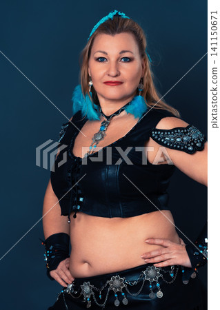 Woman belly dancer wearing leather costume with blue feathers and silver jewelry. Portrait of blonde bellydancer with arms akimbo and looking at camera 141150671
