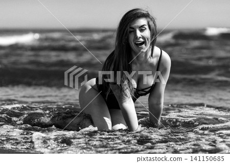Woman laughing in ocean water at beach. Happy lady in swimsuit kneeling in sea waves. Cinematic black and white portrait with film grain and retro aesthetic. Summer vacation and lifestyle concept Woman laughing in ocean water at beach. Happy lady in swimsuit kneeling in sea waves. Cinematic black and white portrait with film grain and retro aesthetic. Summer vacation and lifestyle concept 141150685