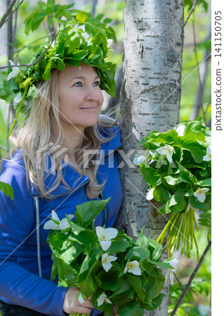 Blonde woman in blue jacket against birch tree holding trillium bouquet wearing leaf crown. Soft natural light filters through spring forest. Serene mood with green foliage white blooms Blonde woman in blue jacket against birch tree holding trillium bouquet wearing leaf crown. Soft natural light filters through spring forest. Serene mood with green foliage white blooms 141150705