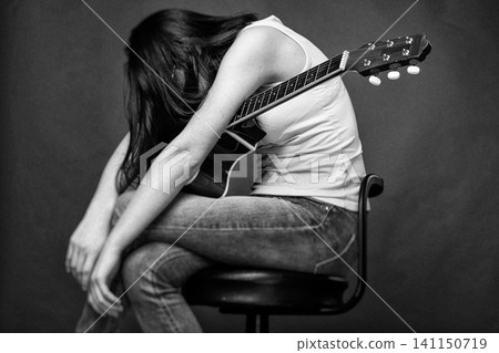 Sad woman hugging guitar on stool. Depressed musician sitting alone with musical instrument. Emotional loneliness and creative crisis. Monochrome portrait in vintage retro style with film grain Sad woman hugging guitar on stool. Depressed musician sitting alone with musical instrument. Emotional loneliness and creative crisis. Monochrome portrait in vintage retro style with film grain 141150719
