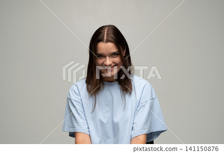 Young woman with brown hair smiling, posing in a light blue t-shirt against a grey background. She is expressing happiness Young woman with brown hair smiling, posing in a light blue t-shirt against a grey background. She is expressing happiness 141150876