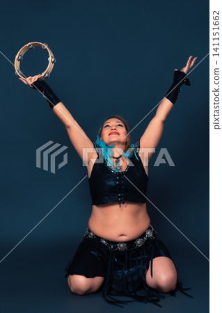 Happy woman dancer is kneeling and raising her arms up, holding tambourine in one of them 141151662