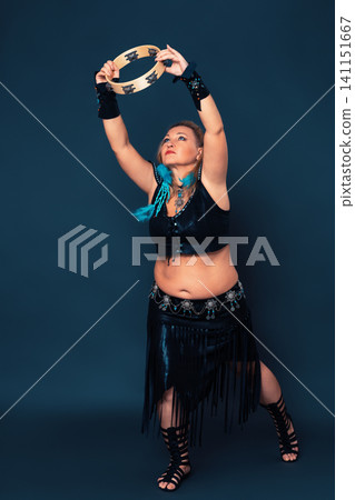 Female dancer in motion holds tambourine above head with both hands and stretches upward in dance Female dancer in motion holds tambourine above head with both hands and stretches upward in dance 141151667