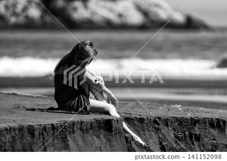 Woman sitting on cliff edge at beach. Lady looking down at sea. Retro style photo, grain effect 141152098