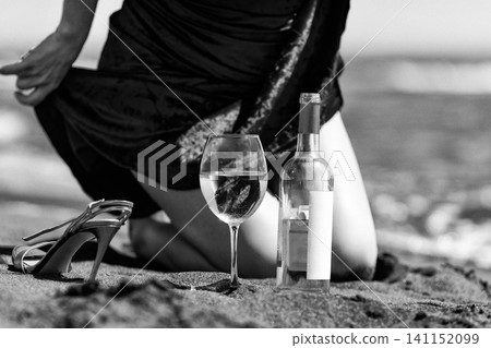 Woman kneeling on sand with wine bottle and glass on beach. Black and white vintage with noise grain Woman kneeling on sand with wine bottle and glass on beach. Black and white vintage with noise grain 141152099