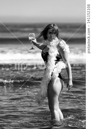 Woman in swimsuit with feather boa holds wine glass in ocean. Retro style beach vacation photography Woman in swimsuit with feather boa holds wine glass in ocean. Retro style beach vacation photography 141152108