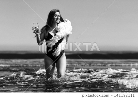 Woman wearing swimsuit and boa stands in ocean holding glass of wine. Monochrome retro style effect Woman wearing swimsuit and boa stands in ocean holding glass of wine. Monochrome retro style effect 141152111