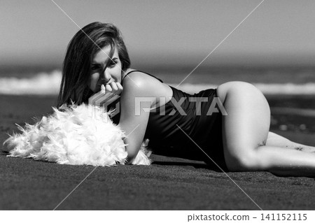 Woman in swimsuit lying on beach sand with white feathers. Monochrome retro style photo of lady Woman in swimsuit lying on beach sand with white feathers. Monochrome retro style photo of lady 141152115