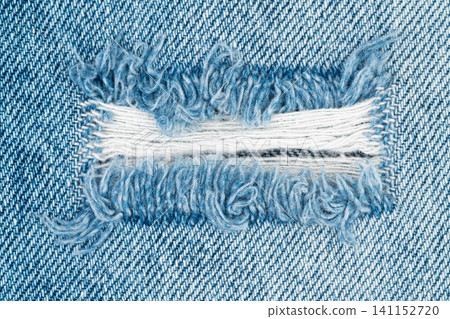 Blue denim fabric with ripped hole feature showing frayed white threads. Distressed jeans texture close up. Material background for fashion apparel design and casual wear concept Blue denim fabric with ripped hole feature showing frayed white threads. Distressed jeans texture close up. Material background for fashion apparel design and casual wear concept 141152720