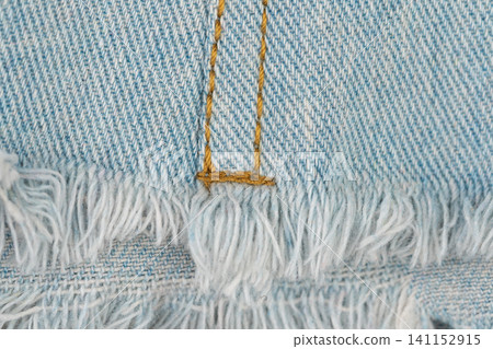 Close-up of light blue denim fabric with distressed fringe edge. Texture detail of casual jeans with detailed stitching. Trendy fashion material for textile background design Close-up of light blue denim fabric with distressed fringe edge. Texture detail of casual jeans with detailed stitching. Trendy fashion material for textile background design 141152915