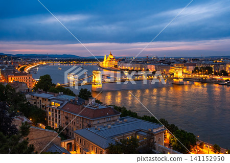 A panoramic view of the blue hour sky and the golden glow of the Danube River at night, from the Royal Castle Hill in Budapest, Hungary. A panoramic view of the blue hour sky and the golden glow of the Danube River at night, from the Royal Castle Hill in Budapest, Hungary. 141152950