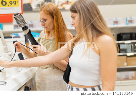 Two women shopping for hair styling tools in a retail store, examining products at a display counter with various items and price tags visible in the background Two women shopping for hair styling tools in a retail store, examining products at a display counter with various items and price tags visible in the background 141153149