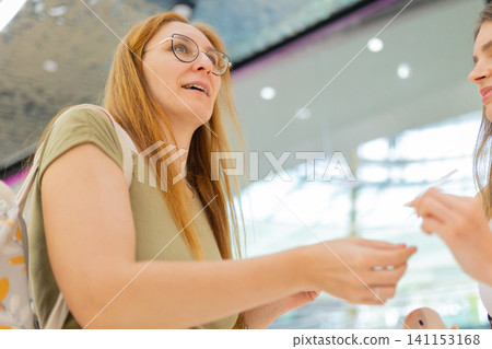 Two women shopping in a mall, one with long hair wearing glasses, interacting while holding shopping bags, bright modern interior with reflective ceiling visible Two women shopping in a mall, one with long hair wearing glasses, interacting while holding shopping bags, bright modern interior with reflective ceiling visible 141153168