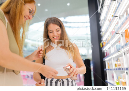 Two women shopping in a mall, examining beauty products on display shelves, with bright lighting and a modern retail environment in the background Two women shopping in a mall, examining beauty products on display shelves, with bright lighting and a modern retail environment in the background 141153169