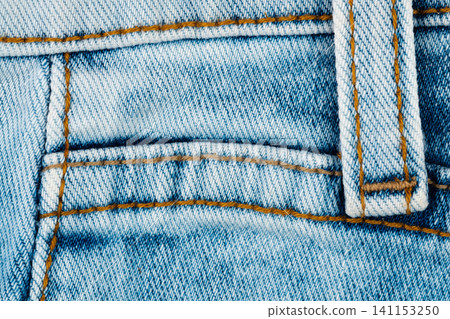Close up of blue denim fabric with gold stitching. Macro detail of jeans waistband and belt loop. Casual clothing material texture for fashion design and textile industry background Close up of blue denim fabric with gold stitching. Macro detail of jeans waistband and belt loop. Casual clothing material texture for fashion design and textile industry background 141153250