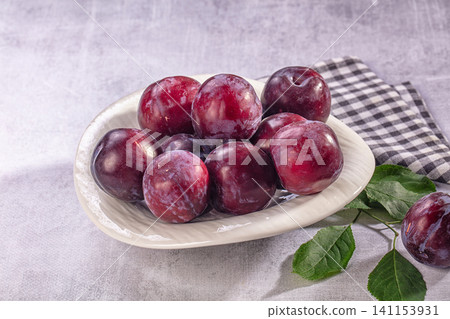 Fresh ripe plums in white ceramic bowl with green leaves and checkered napkin on gray surface Fresh ripe plums in white ceramic bowl with green leaves and checkered napkin on gray surface 141153931