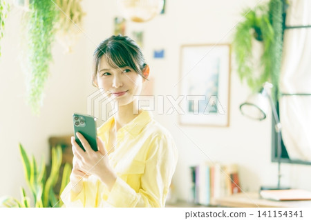 一位年輕的日本女性在擺滿盆栽植物的房間裡使用智慧型手機。 一位年輕的日本女性在擺滿盆栽植物的房間裡使用智慧型手機。 141154341