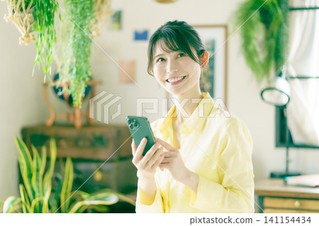 A young Japanese woman smiles as she uses her smartphone in a room surrounded by houseplants. A young Japanese woman smiles as she uses her smartphone in a room surrounded by houseplants. 141154434