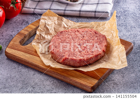 Raw beef patty on parchment paper atop wooden cutting board with tomatoes and kitchen towel in background Raw beef patty on parchment paper atop wooden cutting board with tomatoes and kitchen towel in background 141155504