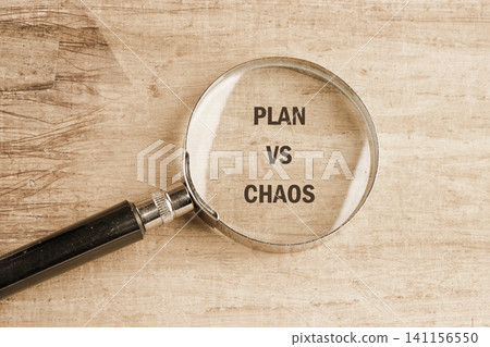 A magnifying glass shows the words plan and chaos on a wooden table in a workspace with some natural light coming in A magnifying glass shows the words plan and chaos on a wooden table in a workspace with some natural light coming in 141156550