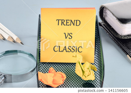 A sticky note with the words trend vs classic is placed on a black tray. Crumpled papers are around it along with pencils and a magnifying glass. Office supplies are in view A sticky note with the words trend vs classic is placed on a black tray. Crumpled papers are around it along with pencils and a magnifying glass. Office supplies are in view 141156605