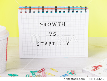 A notepad shows the words growth and stability side by side in large letters. Colorful paper clips lay on the desk near a cup. A bright yellow wall is in the background A notepad shows the words growth and stability side by side in large letters. Colorful paper clips lay on the desk near a cup. A bright yellow wall is in the background 141156642