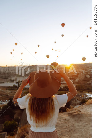 Woman stands on mountain with flying air balloons on background. Tourist Turkish region Cappadocia. Woman stands on mountain with flying air balloons on background. Tourist Turkish region Cappadocia. 141156776
