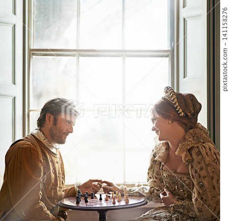 Renaissance, couple and chess date in home with holding hands, vintage and medieval costume in a palace. Window, luxury and queen together with smile from strategy game and talking with love and care Renaissance, couple and chess date in home with holding hands, vintage and medieval costume in a palace. Window, luxury and queen together with smile from strategy game and talking with love and care 141158276