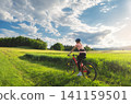 Female cyclist riding mountain bike on path through green fields 141159501