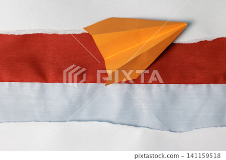 A creative top view composition featuring an orange paper airplane resting on a red and white fabric strip, framed by torn white paper borders. High Quality Photo A creative top view composition featuring an orange paper airplane resting on a red and white fabric strip, framed by torn white paper borders. High Quality Photo 141159518