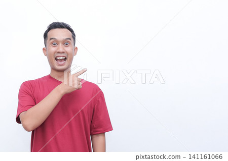 Man in red t-shirt points with wide-eyed surprise. His mouth is open, showing shock or excitement. He gestures toward empty space, drawing viewer's attention. Perfect for advertising 141161066