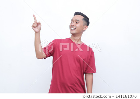 Young man gestures with joy pointing red t-shirt. He smiles warmly, conveying excitement or enthusiasm. Plain white background highlights his expression and motion.Ideal for advertising, presentations Young man gestures with joy pointing red t-shirt. He smiles warmly, conveying excitement or enthusiasm. Plain white background highlights his expression and motion.Ideal for advertising, presentations 141161076