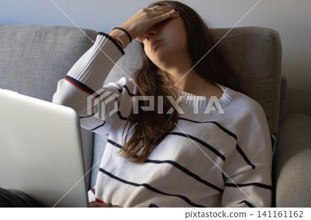 Young woman feeling overwhelmed and stressed from work, covering her face while working remotely on a laptop, struggling with a deadline and experiencing headache and exhaustion 141161162