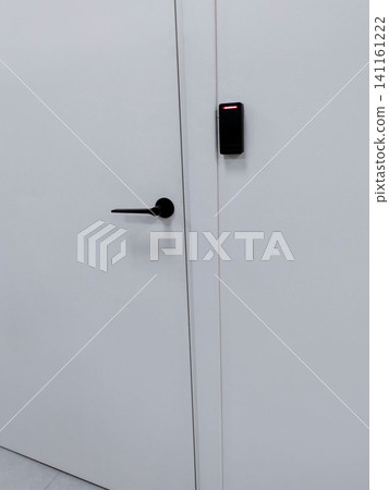 Modern white door with a black handle and electronic access control panel mounted beside it in a minimalist interior setting Modern white door with a black handle and electronic access control panel mounted beside it in a minimalist interior setting 141161222