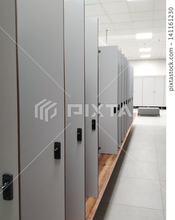 Row of gray lockers with closed doors in a modern changing room, wooden bench visible in the foreground and bright ceiling lights illuminating the space 141161230