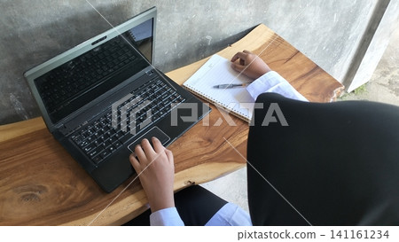 Muslim High School Student Typing on Laptop with Notebook on Wooden Table Muslim High School Student Typing on Laptop with Notebook on Wooden Table 141161234