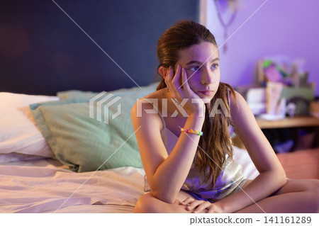 Thoughtful young girl with blue eyes sitting on bed in purple neon light bedroom at night, reflecting on adolescence, emotional journey, and personal growth in a digital age Thoughtful young girl with blue eyes sitting on bed in purple neon light bedroom at night, reflecting on adolescence, emotional journey, and personal growth in a digital age 141161289