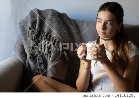 Young woman enjoying a peaceful moment at home, relaxing on a comfortable sofa while holding a warm beverage and contemplating her surroundings Young woman enjoying a peaceful moment at home, relaxing on a comfortable sofa while holding a warm beverage and contemplating her surroundings 141161295