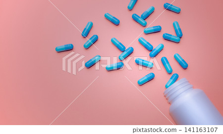 Blue capsule pills spread out of white plastic drug bottle on pink background. Pharmacy banner. Online pharmacy. Painkiller medicine and antibiotic drug resistance concept. Pharmaceutical industry. Blue capsule pills spread out of white plastic drug bottle on pink background. Pharmacy banner. Online pharmacy. Painkiller medicine and antibiotic drug resistance concept. Pharmaceutical industry. 141163107