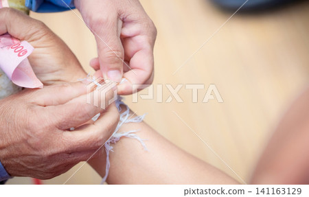The ceremony of binding people wrist with the holy thread to console people's Kwan. Thai culture. Beliefs and local culture of Northeastern Thai people. Old person binding holy thread to Thai adult. The ceremony of binding people wrist with the holy thread to console people's Kwan. Thai culture. Beliefs and local culture of Northeastern Thai people. Old person binding holy thread to Thai adult. 141163129