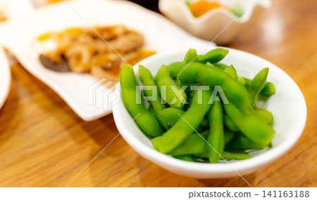 Edamame or green soybeans blanched in white bowl on wooden table at Japanese restaurant. Pods of green soy beans on blurred Japanese food. High fiber and protein food. Plant-based protein. Vegan diet. Edamame or green soybeans blanched in white bowl on wooden table at Japanese restaurant. Pods of green soy beans on blurred Japanese food. High fiber and protein food. Plant-based protein. Vegan diet. 141163188