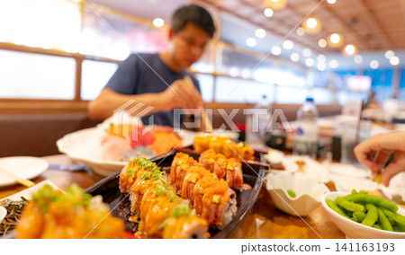 Selective focus on Japanese food in a Japanese restaurant. Salmon sushi on a plate with blurred man eating Japanese food with chopsticks in the restaurant. Healthy Asian food. Salmon sushi menu. Selective focus on Japanese food in a Japanese restaurant. Salmon sushi on a plate with blurred man eating Japanese food with chopsticks in the restaurant. Healthy Asian food. Salmon sushi menu. 141163193