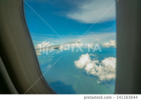 View from inside plane through plane window over blue sky, white clouds and green mountain. Commercial airline flight for summer travel. Plane wing above the city. Airplane flying above land. View from inside plane through plane window over blue sky, white clouds and green mountain. Commercial airline flight for summer travel. Plane wing above the city. Airplane flying above land. 141163644