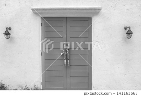 Closed wood door and white concrete wall of the building. Lamp on cement wall near the door. Building exterior. Building wall with rough texture background. Wooden door with handle at the entrance. 141163665
