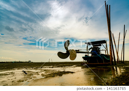 Blade boat on sea coast with blue sky and white clouds at boatyard. Propeller of longtail boat at dock hull. Dry bamboo pole embroidered on the mud beach. Blade boat on sea coast with blue sky and white clouds at boatyard. Propeller of longtail boat at dock hull. Dry bamboo pole embroidered on the mud beach. 141163785