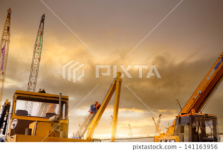 Yellow bulldozer, backhoe, and crawler crane parked at second-hand machinery auction yard with sunset sky and sunlight. Heavy machinery for rent and sale. Crane dealership for a construction business. Yellow bulldozer, backhoe, and crawler crane parked at second-hand machinery auction yard with sunset sky and sunlight. Heavy machinery for rent and sale. Crane dealership for a construction business. 141163956